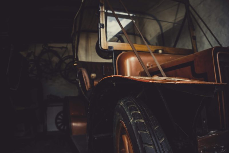 The front of an old vehicle lit warmly in a dark barn, its bonnet and wheel arch catching the light
