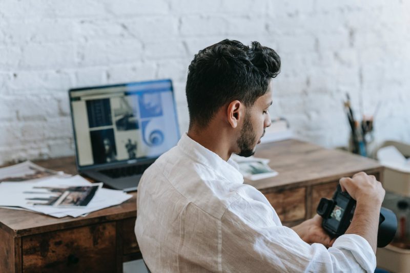 A man at a desk holding a camera, an editing screen and prints in front of him