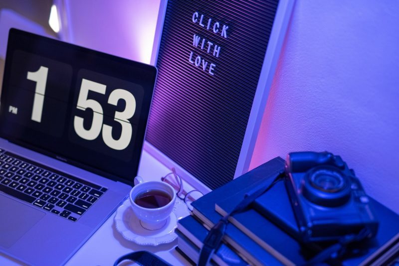 A desk lit purple and blue with a laptop, a clock reading 1:53 and a neon sign
