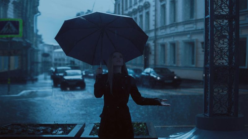 A woman stood with an umbrella outdoors in the rain holding her hand out to feel the rain drops