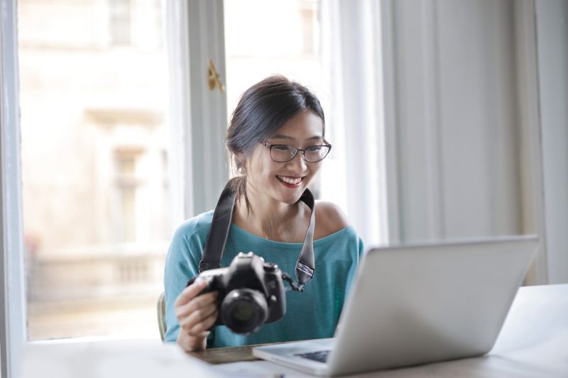A smiling lady wearing glasses with a camera around her neck sat at a table smiling at a laptop
