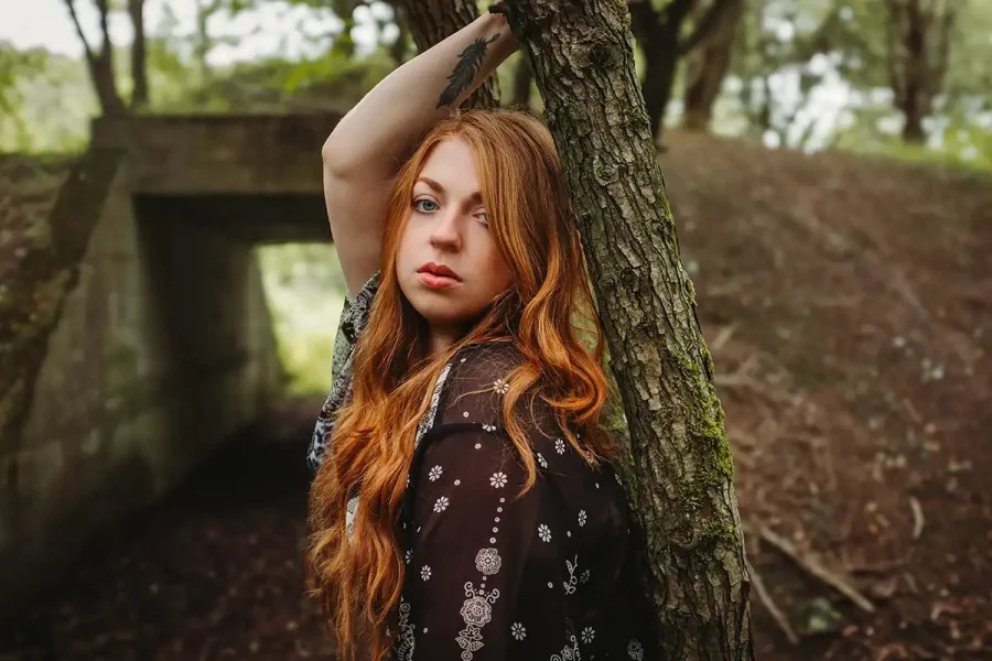 A red-haired woman leaning against a tree, arm raised above her head