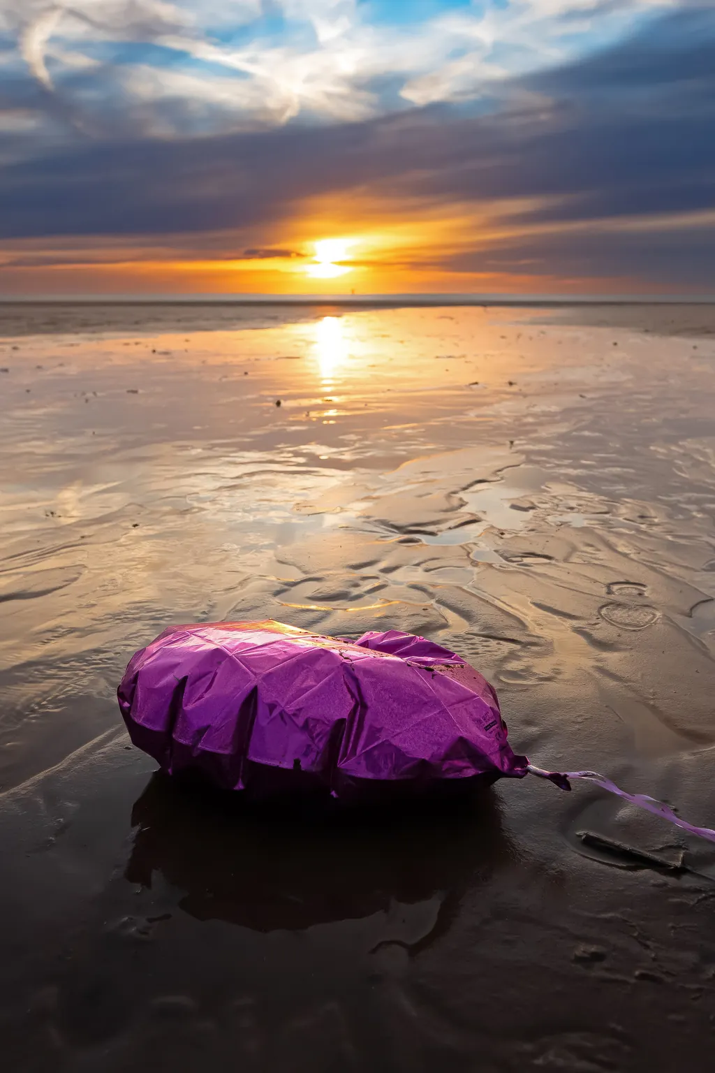 A pink heart-shaped balloon lying on wet sand at sunset