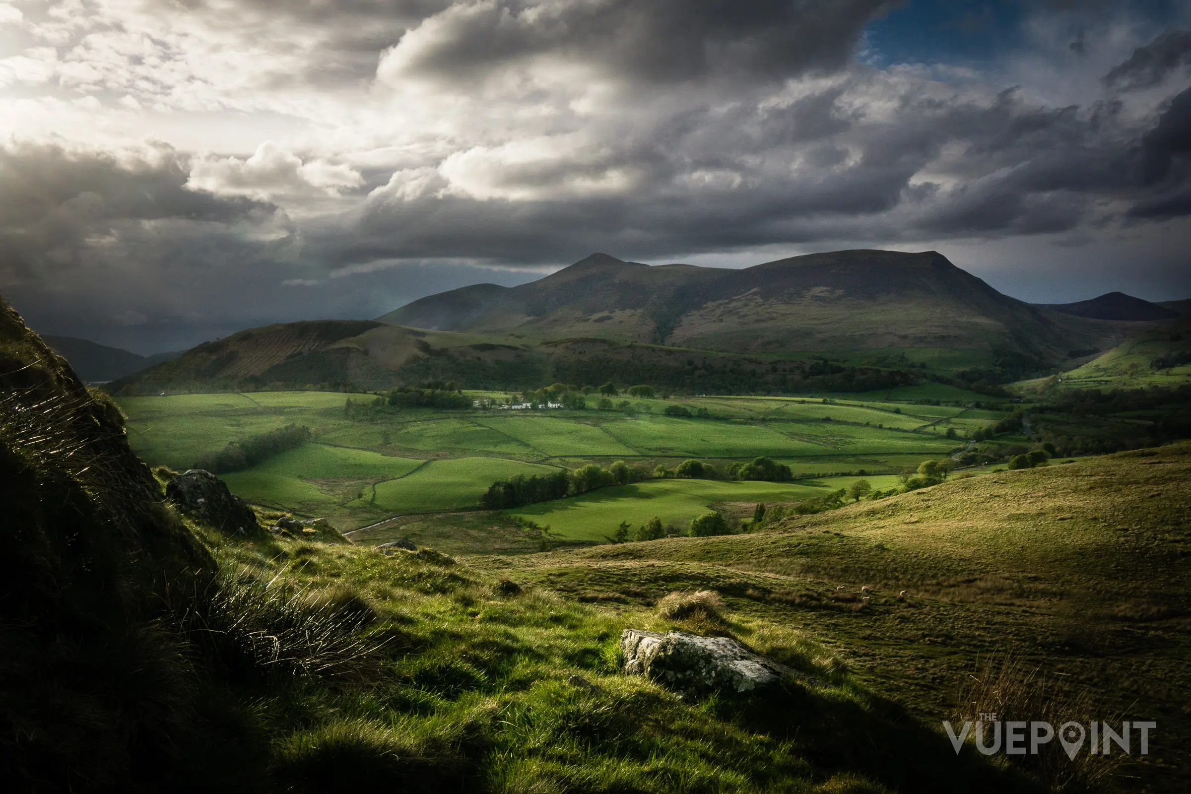 A green valley with mountains behind under a broken sky