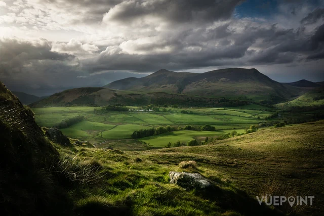 A green valley with mountains behind under a broken sky