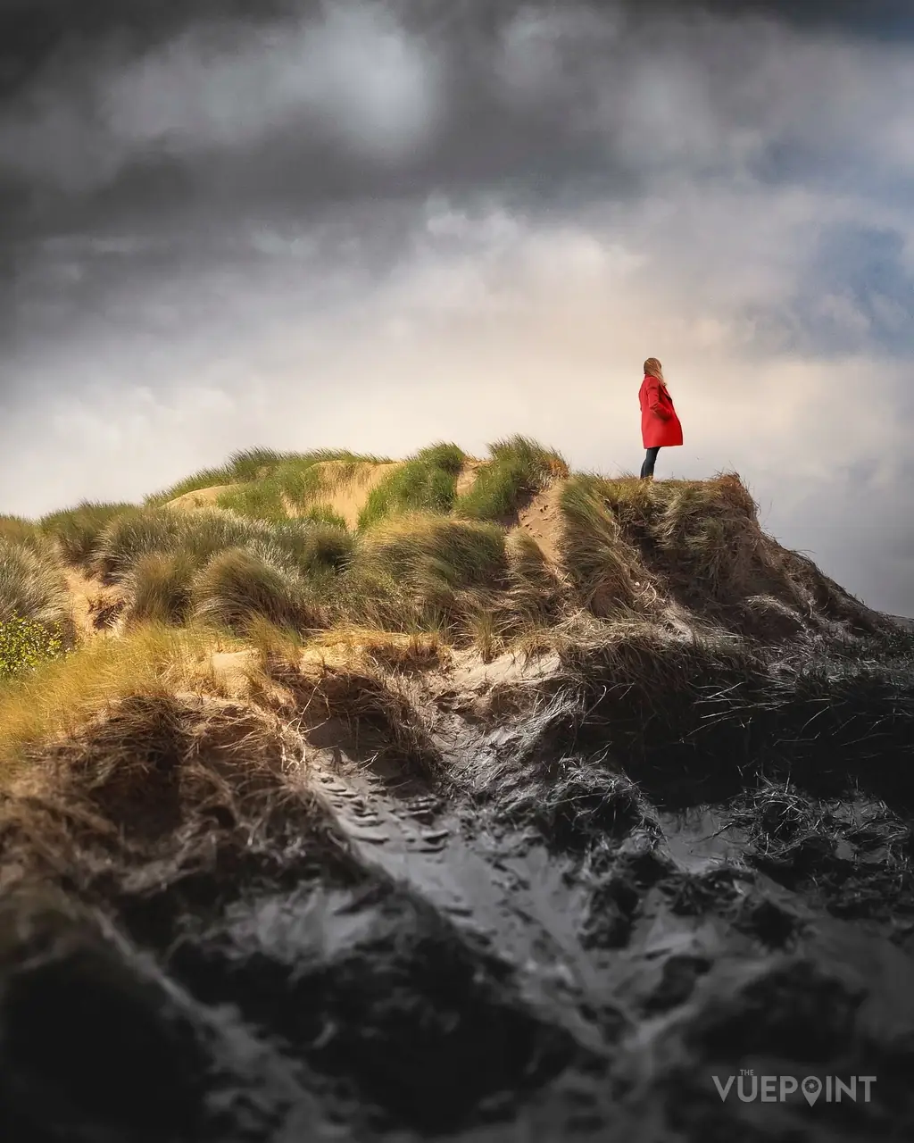 A tiny figure in red standing on a headland above a rough sea