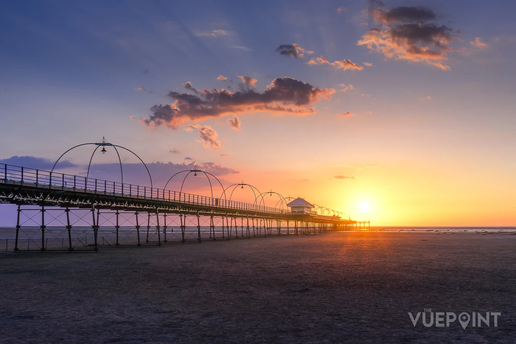Southport Pier in silhouette running out across wet sand, the sun sitting on the horizon beyond the end of it