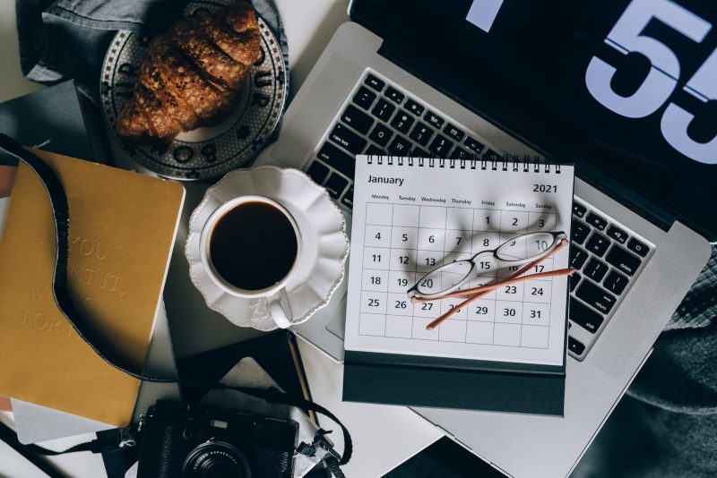Overhead photo of a desk calendar lying on top of a laptop next to a cup of coffee and croissant