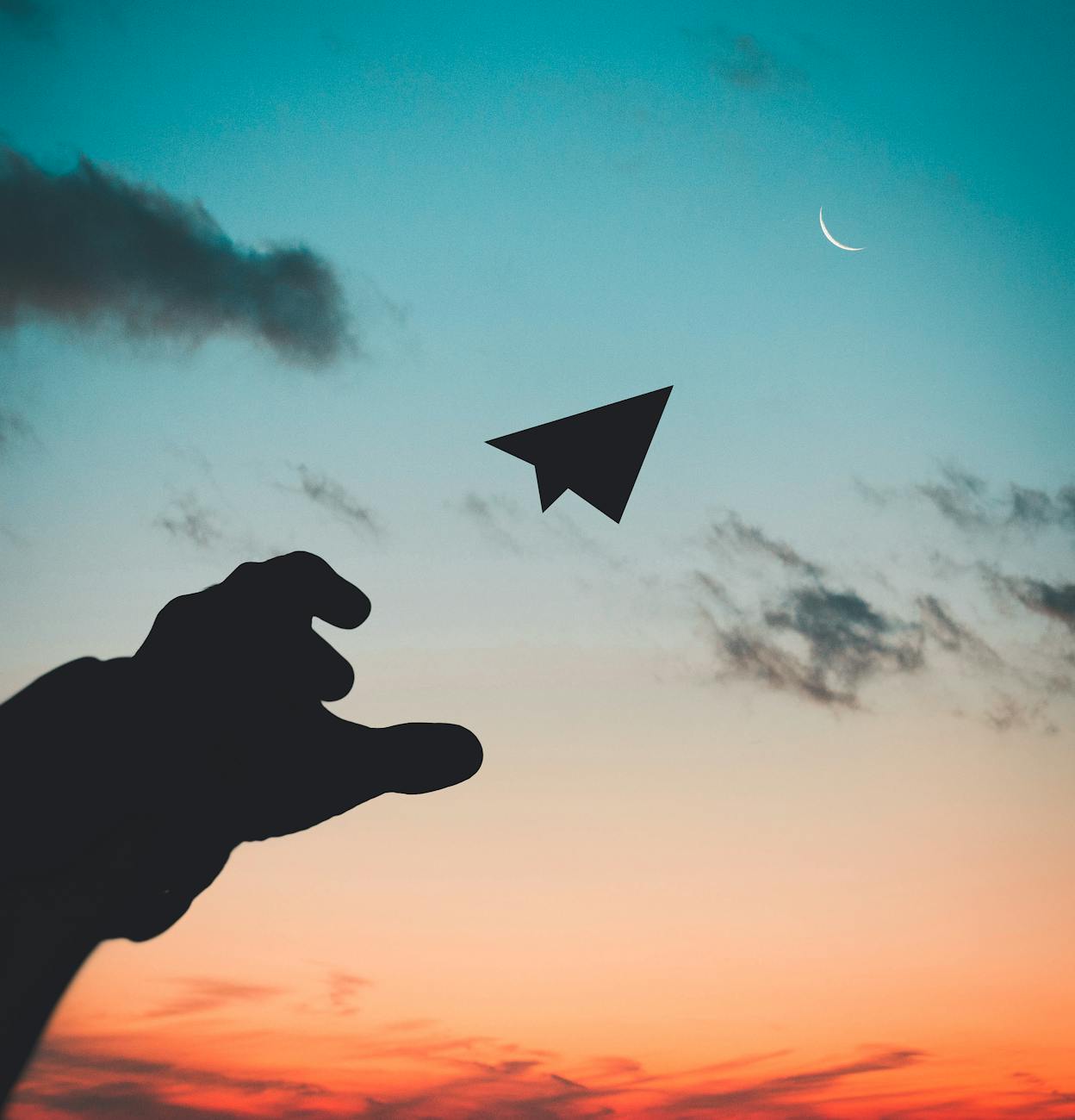 silhouette of a hand throwing a paper airplane towards the moon at sunset