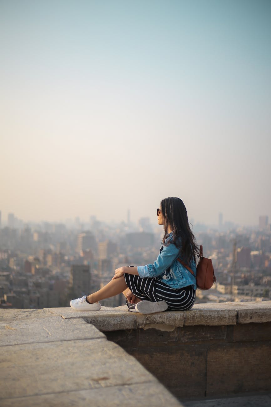 a lady sat on the top of a building overlooking a city. she is wearing a denim jacket and striped dress
