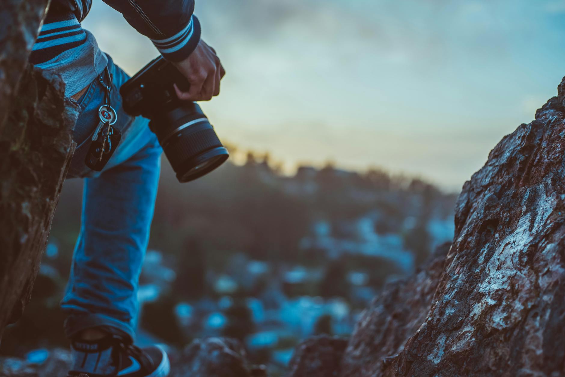 A man sat offscreen holding a camera to his right side on a rock overlooking a landscape
