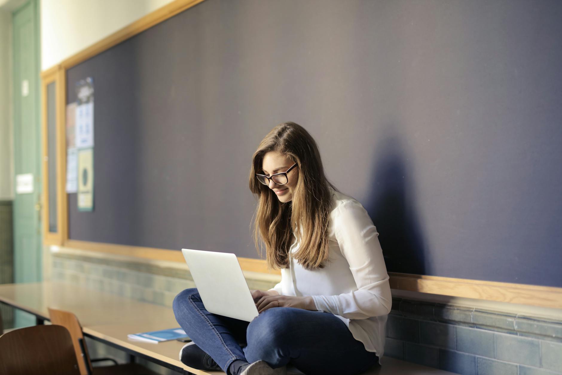 A woman sat crossed leg on a table against a noticeboard with a laptop on her lap smiling and wearing glasses