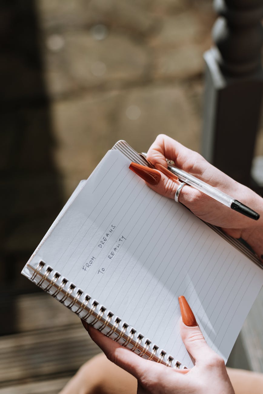 a lady's hands with long orange fingernails holding a notepad which reads from dreams to reality